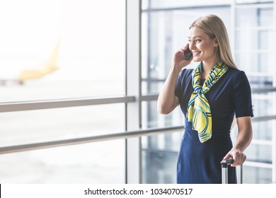 Attractive Young Female Flight Attendant Is Walking In Airport Terminal And Talking On A Smart Phone.