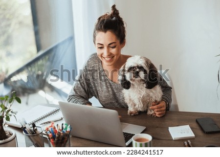 Attractive young female freelancer working on laptop from her home and having her pet dog in her lap to keep her company.