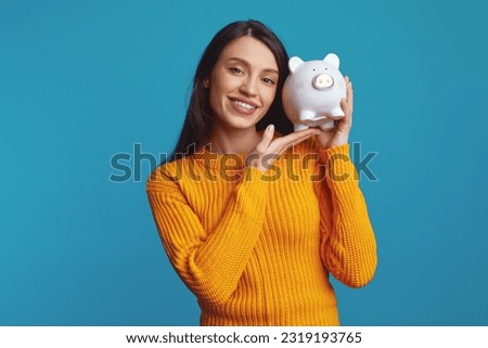 Attractive young female in knitted orange sweater holding white piggy bank with lots of money near face isolated over blue background