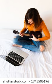 Attractive Young Female Learning To Play Ukelele With Videos On Her Laptop While Sitting On Her Bed