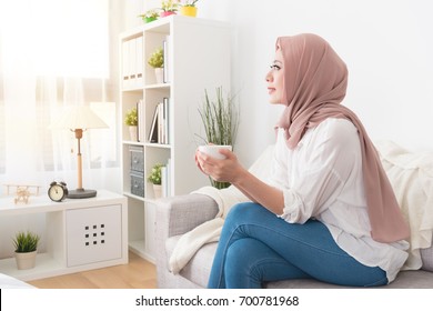 attractive young female muslim holding coffee mug cup sitting on sofa couch and looking at outside daydreaming relaxing enjoying personal afternoon tea time.