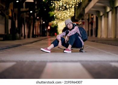 Attractive young female skater in hat and backpack sitting on a skateboard on the night city street. - Powered by Shutterstock