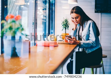 Attractive young female watching video online on smartphone spending free time in cafe interior, hipster girl chatting with boyfriend sending text messages using cheap mobile tariffs for SMS