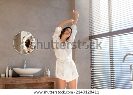 Attractive Young Female In White Silk Robe Relaxing And Stretching In Modern Bathroom, Beautiful Millennial Woman Raising Hands Up And Smiling, Enjoying Morning Time At Home, Copy Space