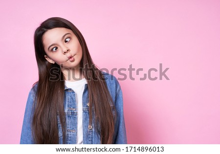 Attractive young funny emotional caucasian girl child long-haired brunette. Squinting, fooling around again in children, copy space. In a white basic t-shirt jeans jacket on a pink background.