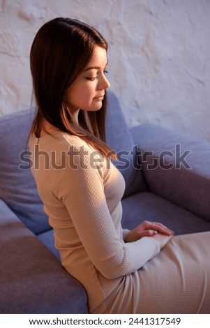 an attractive young girl in a beige casual dress sits relaxing on a purple stylish chair with downcast gaze, a girl with beautiful big eyes with light daytime makeup, clean skin and well-groomed hair.
