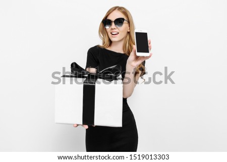 An attractive young girl in a black dress and sunglasses, holding a gift with a black Ribbon, and shows a blank smartphone screen to the camera. Shopping, discounts, online shopping, Black Friday