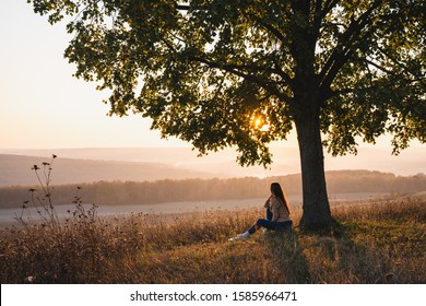 Attractive young girl in casual clothes sitting under big tree with book and looking at the beautiful sunset, autumn sunny day