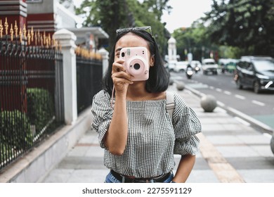 Attractive young girl in casual style taking picture by pink instax camera at the road side