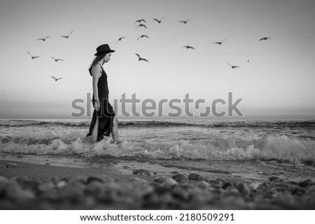 attractive young girl in long dress and hat enters thoughtfully into raging sea. waves evening sea sunset. nostalgia and romantic thoughts. Artistic cinematic look. seagulls in sky. black and white
