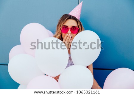 Attractive young girl looks surprised, covering her mouth by hand while standing with air balloons. Wearing pink sunglasses and party hat, outside.