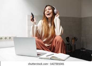 家庭のキッチンにノートパソコンを持って座り、音楽を聴く魅力的な若い女の子