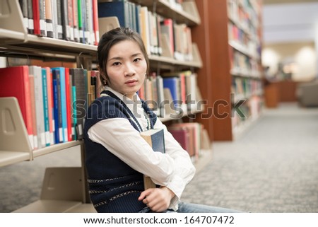 Attractive young girl sitting on the floor in front of a bookshelf holding a thick old book