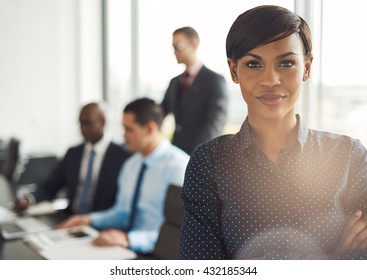 Attractive Young Grinning Business Owner In Office With Polka Dot Blouse, Folded Arms And Confident Expression In Front Of Group Of Employees At Conference Table