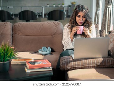 An attractive young happy Indian Asian female or woman sitting on a couch watching an online movie on a Laptop while drinking coffee in an apartment or interior house setup. online entertainment  - Shutterstock ID 2125790087
