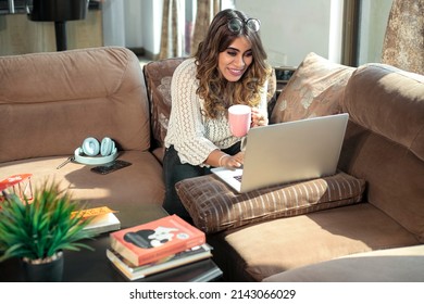 An attractive young happy Indian Asian female or woman sitting on a couch smiling, watching an online movie on a Laptop while drinking coffee in an apartment or interior house. online entertainment 