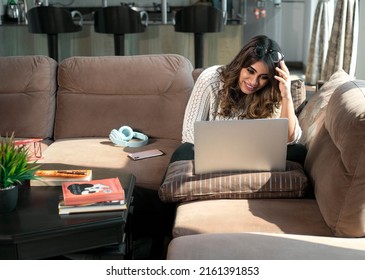 An attractive young happy Indian Asian female or woman with brunette long hair sitting on a couch smiling, watching an online movie on a Laptop in an apartment or interior house. online entertainment - Shutterstock ID 2161391853