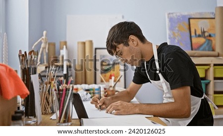 Attractive young hispanic man ardently draws in art studio, engrossed in his smartphone, surrounded by brushes and paint palette