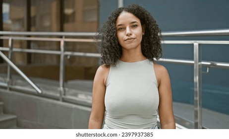 Attractive young hispanic woman, with curly hair, displaying a serious expression while standing on a sunlit city street, living a relaxed yet cool, urban lifestyle. - Powered by Shutterstock