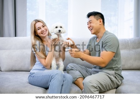 Attractive young husband and wife are sitting on the couch and smiling. They lovingly pet the lapdog