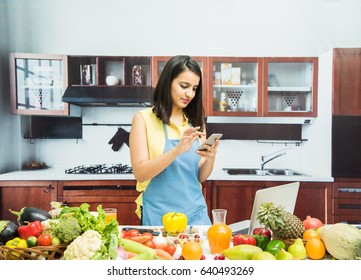 Attractive Young Indian Girl With Apron In Kitchen Using Mobile Phone Or Smartphone With Table Full Of Fruits And Vegetables And Computer