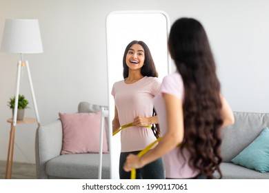 Attractive young Indian woman measuring her waist with tape near mirror indoors, copy space. Young Asian lady happy with results of slimming diet or liposuction, being successful in weight loss - Powered by Shutterstock