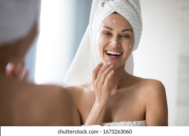 Attractive Young Lady With Towel On Head Applying Moisturizing Smoothing Cream On Face After Showering, Excited By Good Cosmetics Results. Happy Woman Enjoying Morning Skincare Routine In Bathroom.