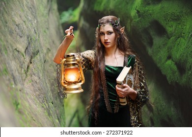 Attractive young long haired brunette female elf walking in the mysterious woods holding a lantern and an old book nature magic mystery cosplay creature mythical romantic fantasy innocence concept.