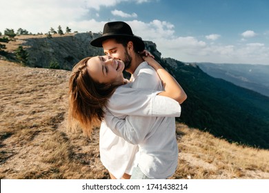 Attractive Young Loving Couple Of Man In White Shirt And Blue Jeans With Girl In Dress On Sunny Outdoor Background In The Green Mountain Landscape