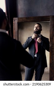 An Attractive Young Man With A Beard Adjusts His Tie In The Mirror