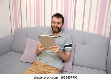 Attractive young man with beard is using a tablet and smiling on sofa at home