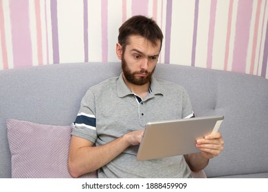 Attractive young man with beard is using a tablet and surprised on sofa at home