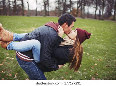 Attractive young man carrying his pretty girlfriend and kissing. Mixed race couple in love outdoors in park.