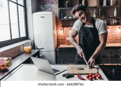 Attractive young man is cooking on kitchen with laptop on table while talking on smart phone. - Powered by Shutterstock