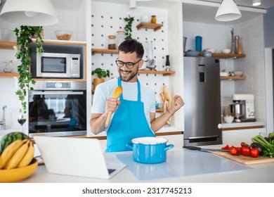 Attractive young man dancing and singing in the kitchen stock photo. happy male holding kitchenware as microphone, listening to music, dancing, doing housework at home, preparing breakfast - Powered by Shutterstock