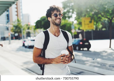 Attractive young man with dark curly hair wearing sunglasses is walking the street with a cup of coffee on a sunny day. Bearded hipster guy wearing casual clothes enjoys relaxing on a summer weekend.