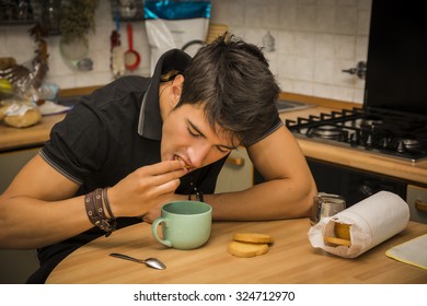 Attractive Young Man Eating Breakfast, Leaning With Elbow On Kitchen Table And Holding Cup Of Coffee For Breakfast