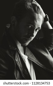 Attractive young man with fair hair wearing stud, leather jacket and v-neck t-shirt, holding one hand in his hair, posing in a dark room next to the window. Looking at camera.Black and white.Texture.