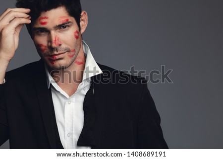 Attractive young man full of kiss marks on his face. Man in love against grey background.