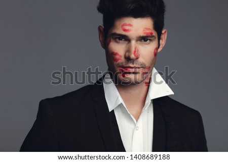 Attractive young man full of lipstick marks of kisses on his face. Handsome male model in black blazer standing against grey background.