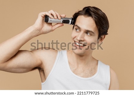 Attractive young man he 20s perfect skin in undershirt hold electric razor grooming with trimmer look aside isolated on plain pastel beige background. Skin care healthcare cosmetic procedures concept