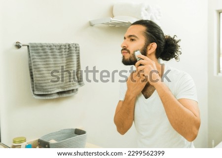 Attractive young man in his 20s using a trimmer and grooming his beard in the bathroom mirror