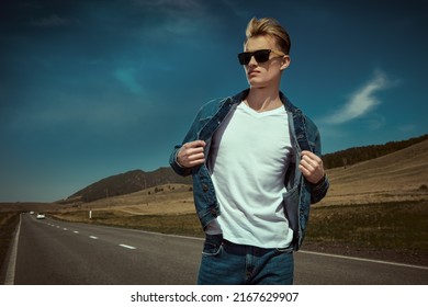 Attractive Young Man Hitchhiker In Denim Clothes And Black Sunglasses Stands On The Road On The Background Of Picturesque Landscape. Denim Fashion. Road Adventures, Travelling.