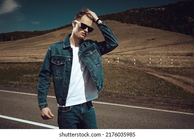 Attractive Young Man Hitchhiker In Denim Clothes And Black Sunglasses Stands On The Road On The Background Of Picturesque Landscape. Denim Fashion. Road Adventures, Travelling.