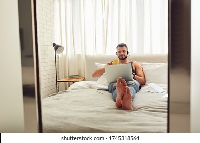 Attractive Young Man Holding Cup Of Hot Drink And Watching Movie On Notebook While Resting In Bedroom