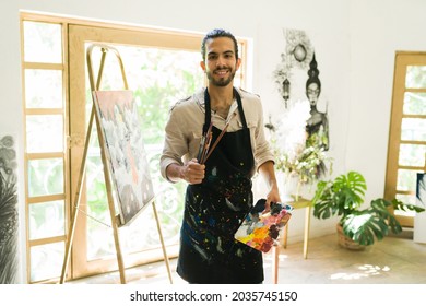 Attractive young man holding his paint brushes and a palette ready to create an artistic painting at the studio