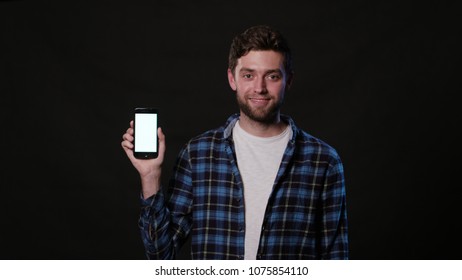 An attractive young man holding a phone with a white screen against a black background. Medium Shot