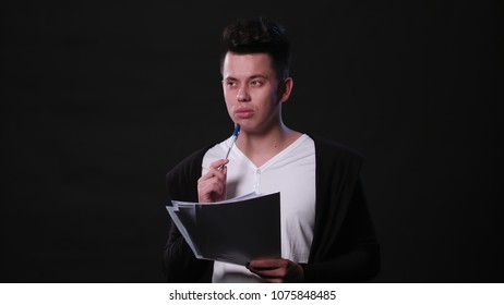 An attractive young man holding sheets of paper and writing against a black background. Medium Shot