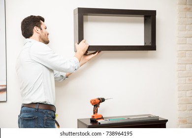 Attractive Young Man Installing A Shelf On A Wall Using A Power Drill And A Level