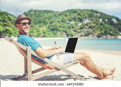 Attractive Young Man With Laptop Working On The Beach. Freedom, Remote Work, Freelancer, Technology, Internet, Travel And Vacation Concepts